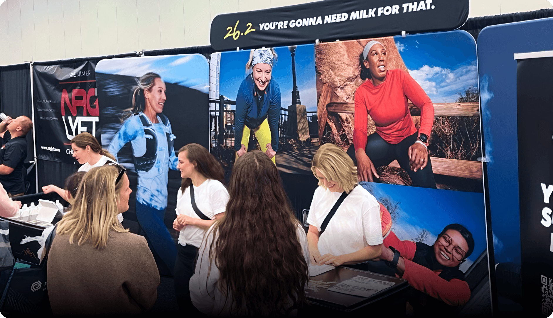 An expo booth with banner ‘26.2 You’re Gonna Need Milk for That’ surrounded by display images of female runners.