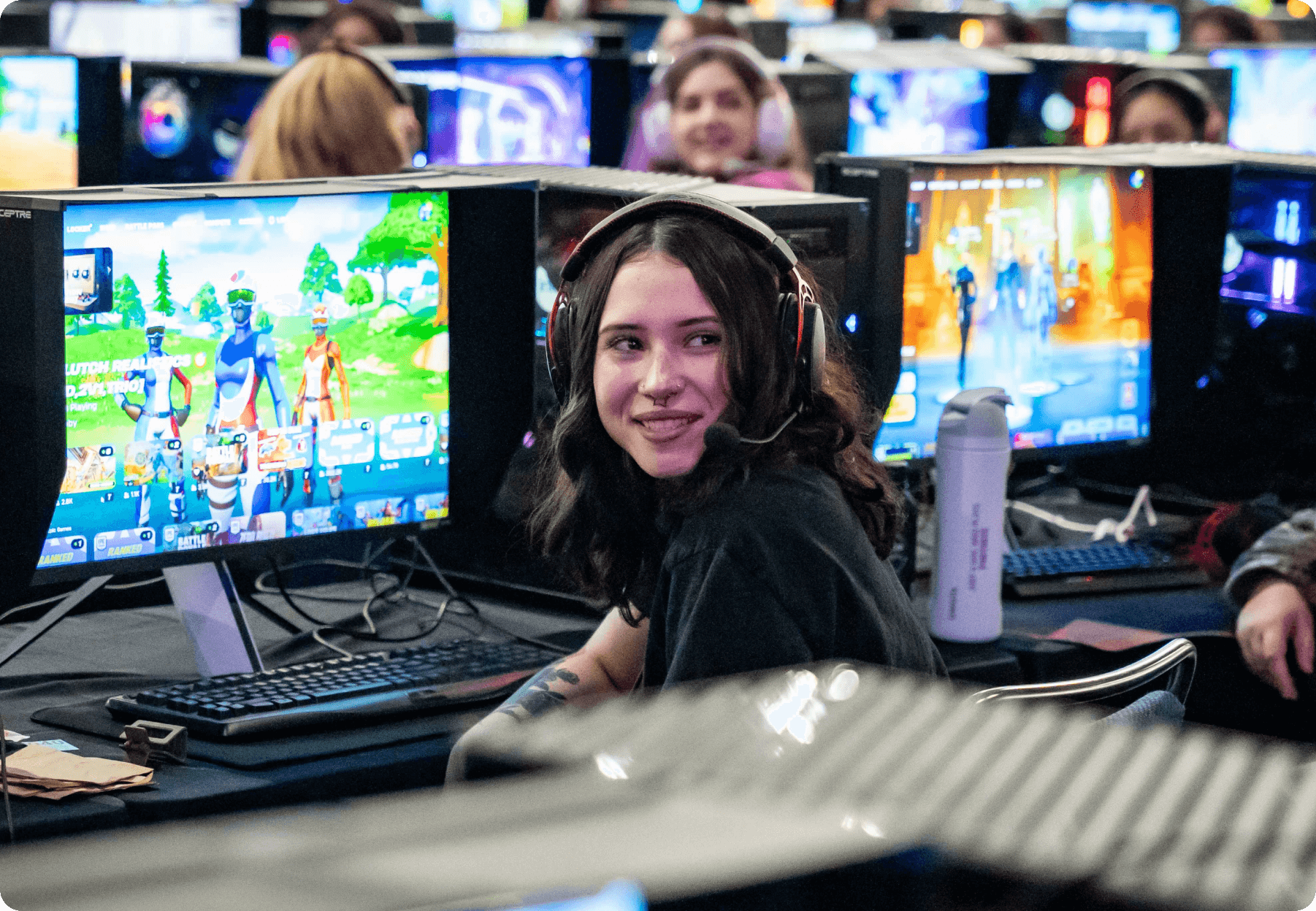 A woman playing at a local Fortnite scrimmage, wearing headphones and  surrounded by other female gamers.