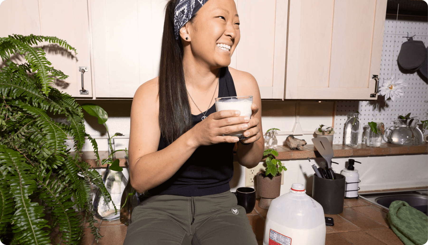 A female sits on her kitchen counter drinking a glass of white milk, a gallon of whole milk sits on the counter, next to a variety of plants.