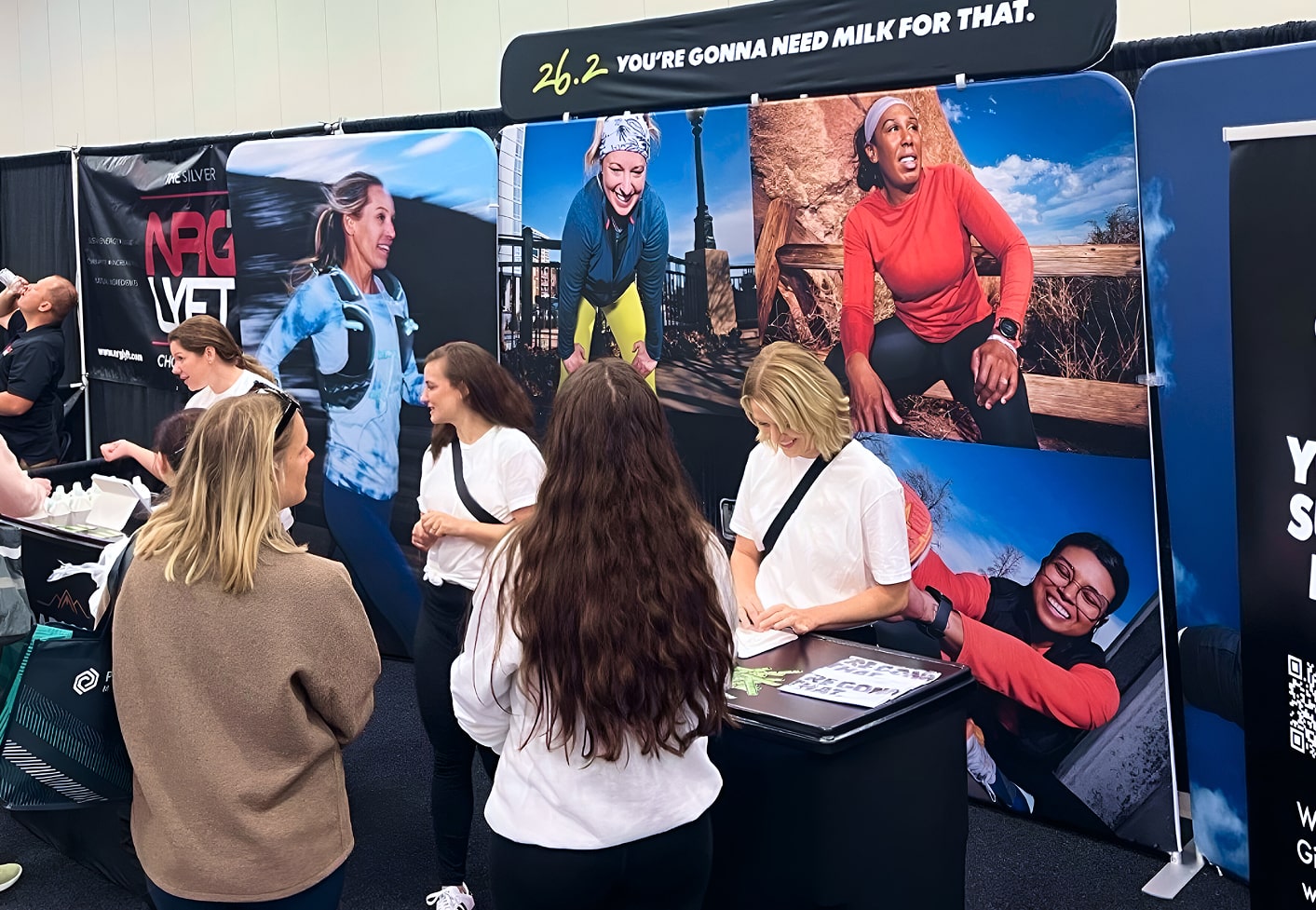 An expo booth with banner ‘26.2 You’re Gonna Need Milk for That’ surrounded by display images of female runners.